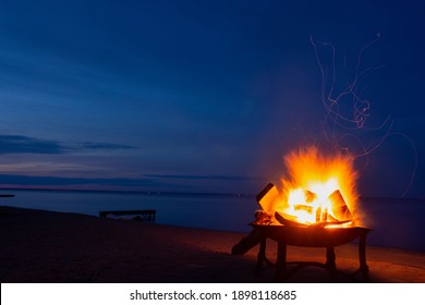 Burning Fire In A Fire Pit And Wooden Dock On The Beach Facing Lake Saint-Jean In Quebec, Canada At Dusk On A Warm Summer Night