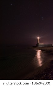 A burning lighthouse by the sea at night illuminates the path. Glowing lighthouse against the background of the night sky and stars. A lighthouse indicates the path to the sea. Night view