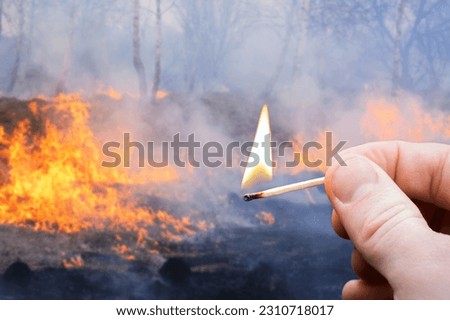 Burning match in hand of a man and flames of wildfire spreading through the forests and fields on the background. Wildfire concept, small match causes fire, disastrous consequences for nature, people