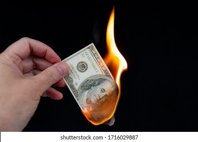 Burning a US $100 bill. A hand holds the burning banknote, about half of it has been burned. Black background.