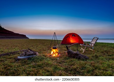 Burning Wood Bonfire With Gentle Flames And Red Tent, Leisure Chair By The Lake On A Sunny Sunset.