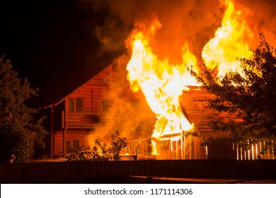 Burning wooden house at night. Bright orange flames and dense smoke from under the tiled roof on dark sky, trees silhouettes and residential neighbor cottage background. Disaster and danger concept. - Powered by Shutterstock