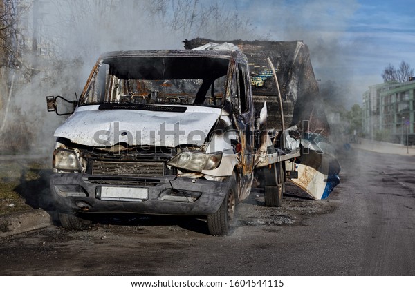 Burnt out city truck car by fire accident at the\
road side in the city. Burned out wrecked damaged car with smoke\
clouds after street protests, mass riots, arson. Car fire accident\
insurance concept