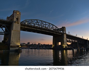 Burrard Street Bridge in the evening light after sunset with illuminated pillars reflected in the smooth water of False Creek bay in Vancouver, British Columbia, Canada with orange colored sky.