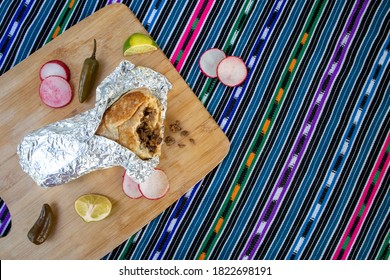 Burrito Wrapped In Foil Surrounded By Peppers, Radishes, And Lime On A Wood Surface On A Colorful Tablecloth - Overhead