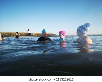 Burry Port, Wales, UK: January 13, 2022: A Small Group Of Ladies Swim In Open Sea Water During The Winter Months For Physical And Mental Health Benefits. They Are Wearing Wetsuits And Bobble Hats To 