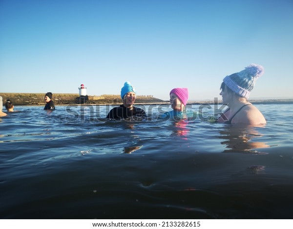 Burry Port, Wales, UK: January 13, 2022: A small group
of ladies swim in open sea water during the winter months for
physical and mental health benefits. They are wearing wetsuits and
bobble hats to 