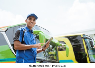 A Bus Crew Member In Uniform And A Cap Smiling At The Camera While Using A Digital Tablet Against The Background Of The Bus Fleet