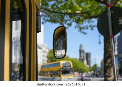 Bus mirror with Berlin backdrop. Side view of a yellow bus with a focus on the exterior mirror, which reflects the Berlin cityscape. In the background, a double-decker bus and the Kaiser Wilhelm Memor