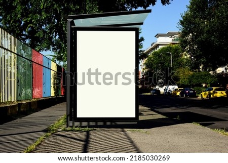 bus shelter blank ad panel. billboard display. empty white lightbox sign at busstop. glass and aluminum structure. city transit station. urban street and green park setting. outdoor advertising.