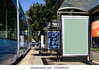 Bus Shelter With Empty White Ad Panel And Light Box. Urban Setting With Green Park. Billboard For Mock-up, Advertising Background. Place Holder Sample Image. Glass And Stainless Steel Structure.