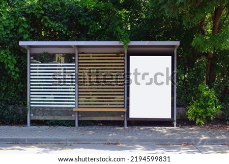 bus shelter with empty white ad panel and lightbox. billboard for mockup and advertising background. blank copy space placeholder. glass and aluminum structure. wood bench. green foliage background. 