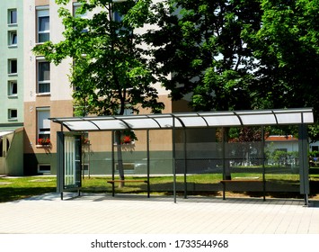 Bus Shelter And Bus Stop Of Clear Glass And Aluminum Frame Structure In Residential Area. Green Trees And Lawn In The Background. Public Transportation. Ad And Billboard Space On The Sides.