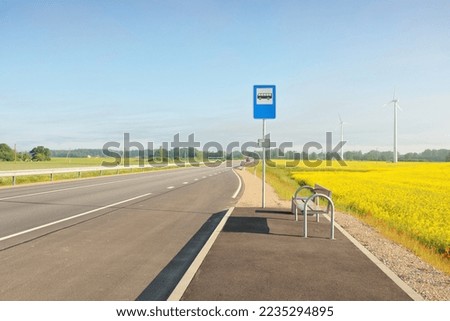 Bus stop and country highway (new asphalt road) through the green forest meadow and blooming yellow rapeseed field at sunrise. Fog, soft sunlight. Transportation, remote places, vacations concepts