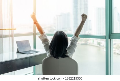 Business achievement concept with happy businesswoman relaxing in office or hotel room, resting and raising fists with ambition looking forward to city building urban scene through glass window