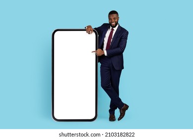 Business Advertisement. Smiling African American Businessman Pointing At Big Blank Smartphone With White Screen While Standing On Blue Studio Background, Male Entrepreneur Showing Copy Space, Mockup
