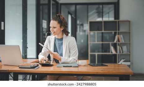 Business analytics concept, Cheerful businesswoman organizing her schedule with a notebook while sitting at a wooden desk with a laptop in a contemporary office.

 - Powered by Shutterstock