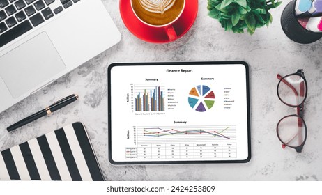 Business analytics concept, A tablet showcasing a finance report with colorful graphs beside a red coffee cup, laptop, and stationery on a marble background.