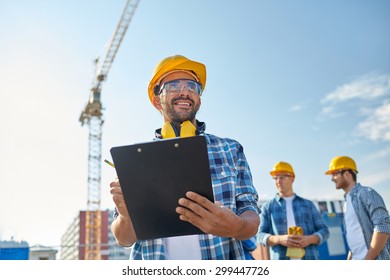 Business, Building, Paperwork And People Concept - Happy Builder In Hardhat With Clipboard And Pencil Over Group Of Builders At Construction Site