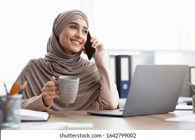 Business Call. Happy arabic woman talking on mobile phone in office and holding cup of coffee, having break at workplace, copy space