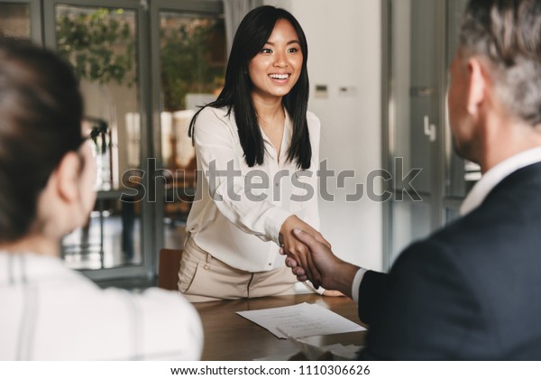 Business, career and
placement concept - image from back of two employers sitting in
office and shaking hand of young asian woman after successful
negotiations or
interview