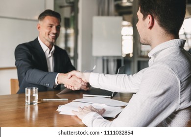 Business, career and placement concept - successful young man smiling and handshaking with european businessman after successful negotiations or interview in office