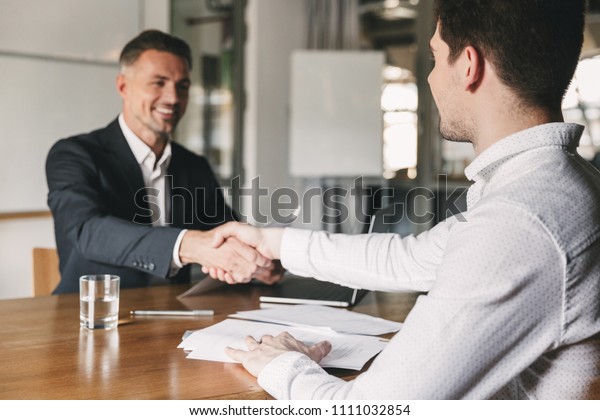 Business, career and placement\
concept - successful young man smiling and handshaking with\
european businessman after successful negotiations or interview in\
office