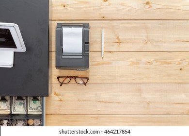 Business Cash Register On A Clean Wooden Desk Or Counter With An Open Cash Drawer Showing American Currency And A Pair Of Folded Spectacles, Overhead View With Copy Space