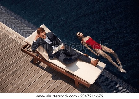 Business clothes, reading newspaper. Man with woman is in the pool together.