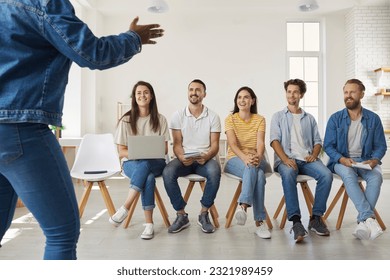 Business coach standing and speaking in front of team of interested people. Group of happy men and women sitting in row on chairs and listening to female speaker sharing her knowledge and experience - Shutterstock ID 2321989459