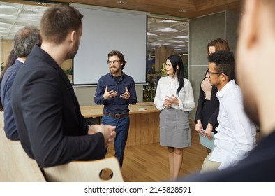 Business Coach Talking To Group Of People Standing In Office Around Him. Team Of Entrepreneurs And Listening To Business Trainer Speaking About Creativity And Teamwork, Sharing Advice And Experience