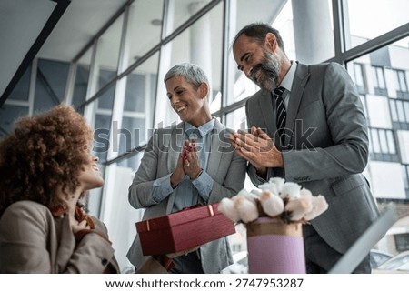 Business colleagues clapping and smiling while presenting a red gift box to a happy female employee, celebrating her achievements and showing appreciation in the office