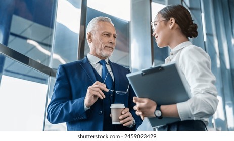 Business colleagues discuss business documents standing indoors. Two diverse partners walking and discussing project in office. Professional business people working together at workspace. - Powered by Shutterstock