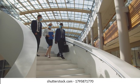 Business Colleagues Walking Downstairs And Chatting Wearing Safety Masks. Diverse Employees In Medical Mask Descending Staircase And Discussing Project In Office Building