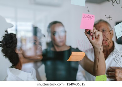 Business Colleagues Working On Project Together. Business Woman Writing On Sticky Note On Glass Wall With Coworkers Standing By In Office.