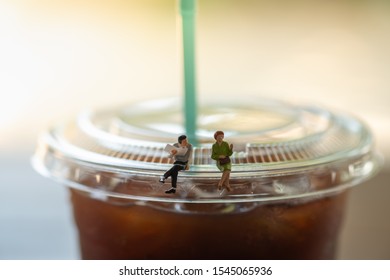 Business And Communication Concept. Businessman With Newspaper And Woman Miniature Figures Sitting And Talking On Top Of Cover Lid Of Plastic Cup Of Iced Black Coffee (Americano)