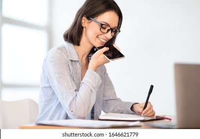 Business Communication Concept. Cheerful boss woman using voice command recorder on smartphone and taking notes