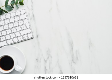 Business composition with white computer keyboard, cup of coffee and eucalyptus plant on white marble background. Concept of coffee break at the office. Top view with copy space for text.