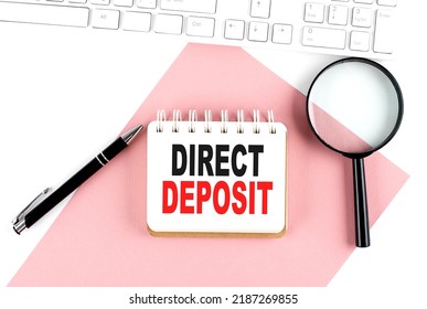 Business Concept. Notebook With Text DIRECT DEPOSIT On Pink Paper With Magnifier, Keyboard And Pen In White Background