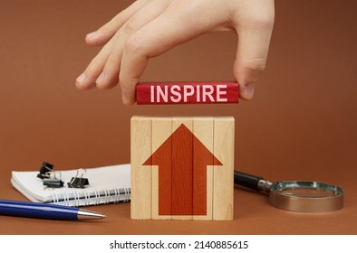 Business Concept. On A Brown Surface Are Office Items, Wooden Blocks, In The Hand Is A Red Block With The Inscription - Inspire