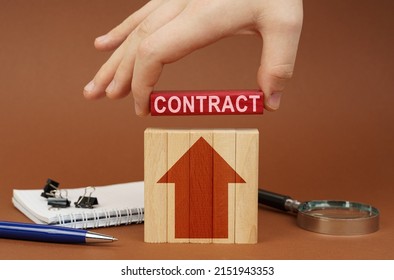 Business Concept. On A Brown Surface Are Office Items, Wooden Blocks, In The Hand Is A Red Block With The Inscription - CONTRACT