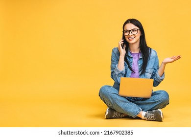 Business concept. Portrait of happy young woman in casual sitting on floor in lotus pose and holding laptop isolated over yellow background. Using mobile phone.