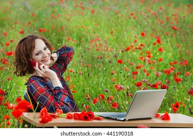 Business Concept Shot Of A Beautiful Young Woman Sitting At A Desk Using A Computer In A Poppy Field. Young Businesswoman In Sunny Poppy Nature Office. Young Woman In Red Dress With Laptop Outside.