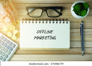 Business Concept - Top view of calculator, coins, jar, eye glasses plant, pen and open notebook written with OFFLINE MARKETING on wooden background.