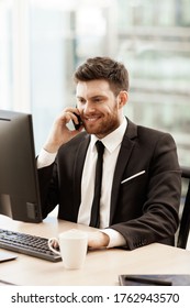 Business Concept. Young Businessman Sitting At The Office Table Happy Talking On A Cell Phone Getting Good News About His Work. Man In Suit Indoors On Glass Window Background.