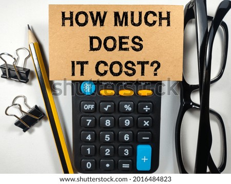 Business concept.Text HOW MUCH DOES IT COST? writing on brown card with calculator,pencil,paper clips and glasses on white background.
