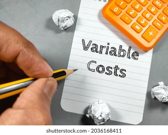Business concept.Text Variable Cost writing on notepaper with hand holding pencil,torn paper  and calculator on gray background.
