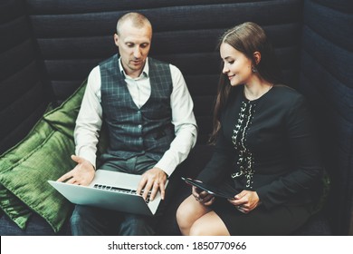 A business couple discussing a new startup project: a businesswoman with a digital tablet is looking on a screen of a laptop of a man entrepreneur sitting next to her in an office coworking sofa - Powered by Shutterstock