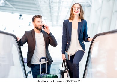 Business Couple Talking With Phones While Getting Up On The Escalators With Baggage At The Airport