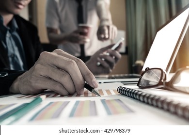business documents on office table with smart phone and laptop and two colleagues discussing data in the black and white background 